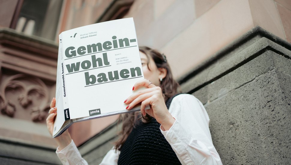 An eine Wand gelehnt liest eine Frau „Gemeinwohl bauen, Band 2: BOB CAMPUS – von der Magie des Gemeinsinns“