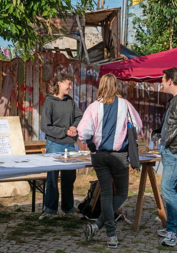 Zwei Personen, die mit einer Frau an einem Stand reden.