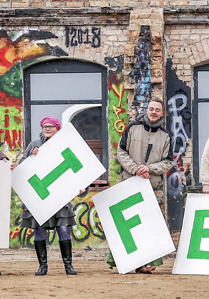 Acht Menschen, die Plakate mit den einzelnen Buchstaben hochhalten, die “FreiFeld” bilden.