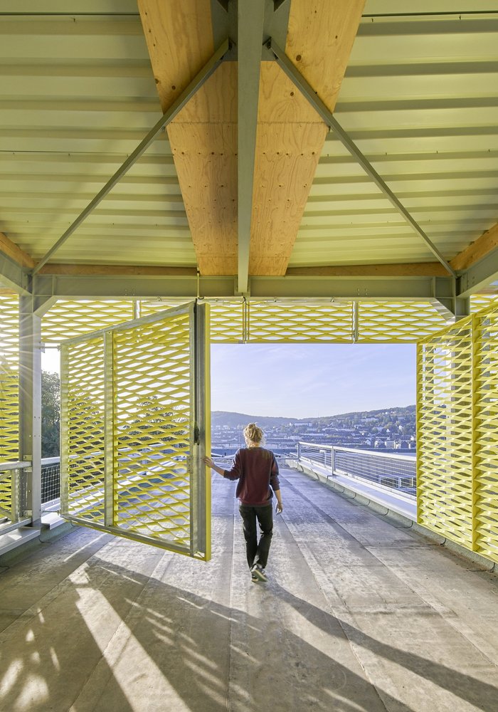 Die Dachterrasse des BOB CAMPUS, die zum Teil mit einem gelben Stahlgerüst überbaut ist. Eine Frau öffnet die Türe zum freien Bereich.