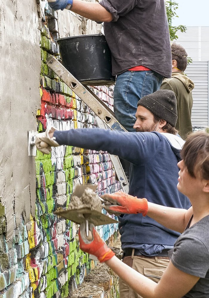 Mehrere Menschen verputzen eine Wand, welche mit Graffiti bemalt ist.