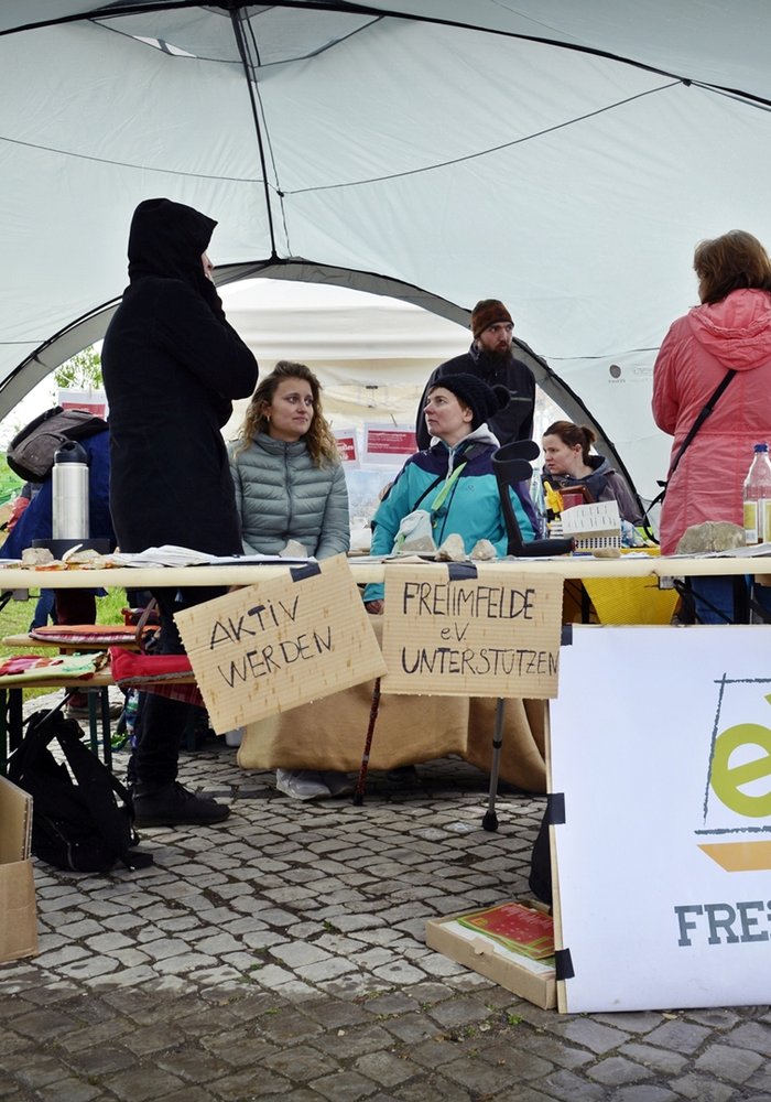 Ein Stand des FreiimFelde e.V. an dem Menschen sitzen mit Schildern auf denen „Aktiv werden“ und „Freifeld unterstützen“ steht.