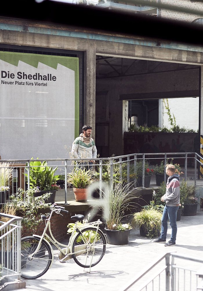 Der Innenhof mit Blick auf die Schedhalle, in dem viele Pflanzen stehen und zwei Menschen, die sich unterhalten