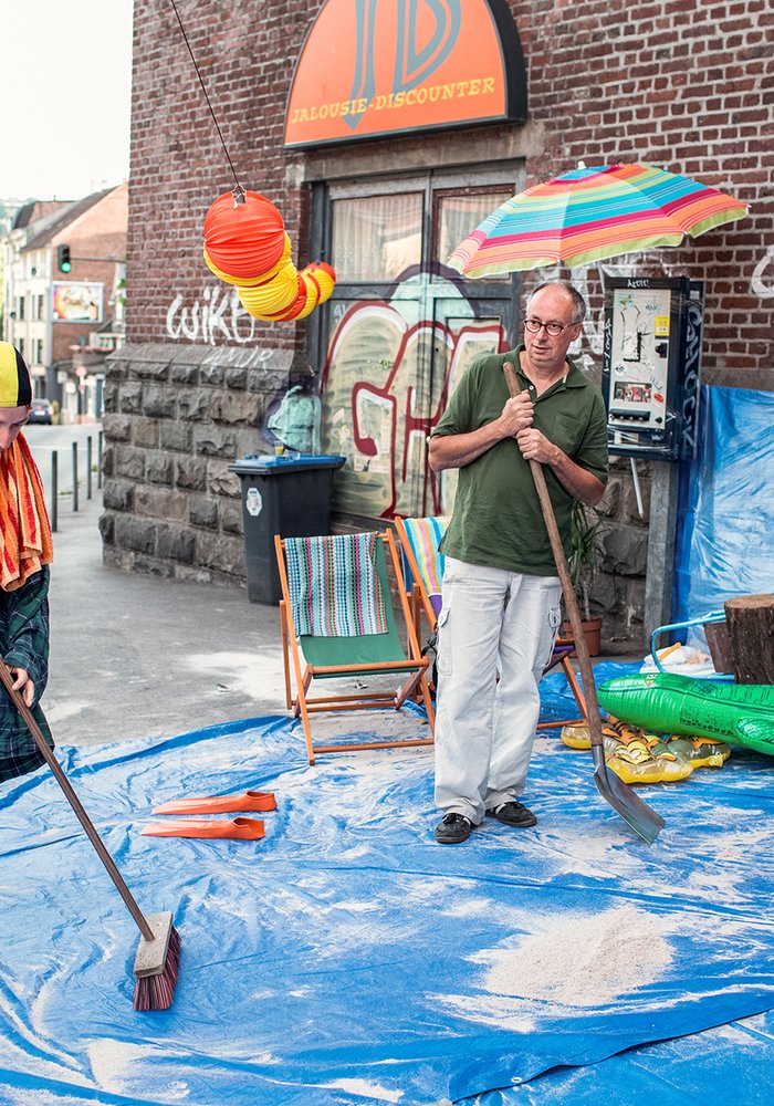Mehrere Menschen kehren auf einer Plastikplane, um die Stranddeko steht, Sand zusammen. Einer der Personen ist verkleidet.