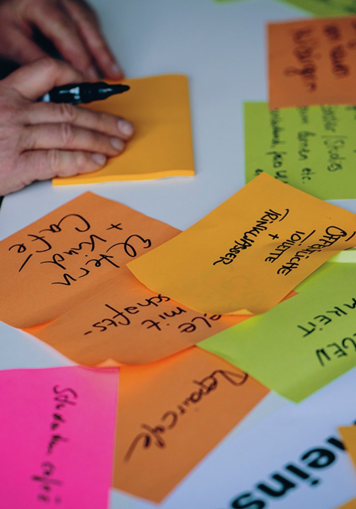 Mehrere beschriebene Post Its, welche auf einem Blatt kleben. Eine Person beschriftet ein weiteres.