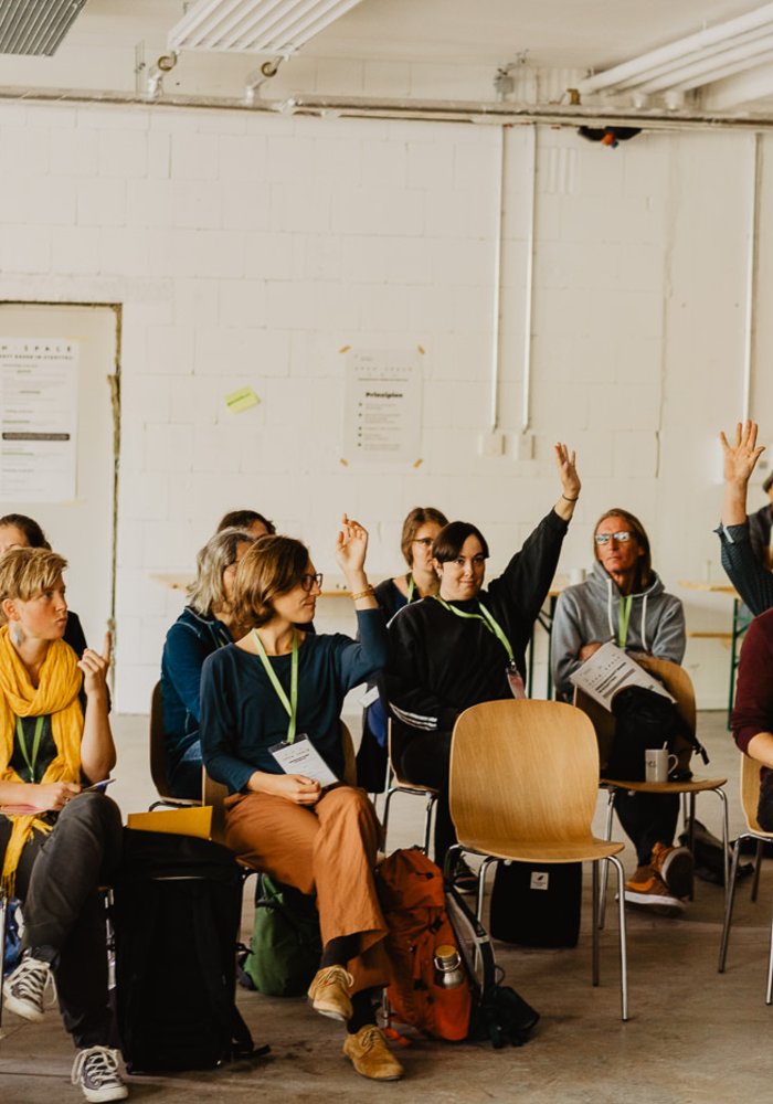 Drei Stuhlreihen, auf denen Menschen sitzen, die sich melden.