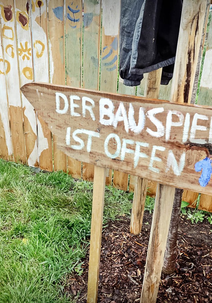 Ein bemalter Holzzaun, welcher von einem Kind weiter bemalt wird. Davor steht ein Holzschild mit der Beschriftung “Der Bauspielplatz ist offen”.