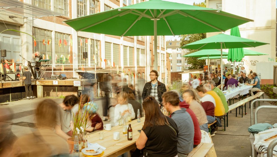 Viele Menschen sitzen an langen Tischen unter grünen Schirmen
