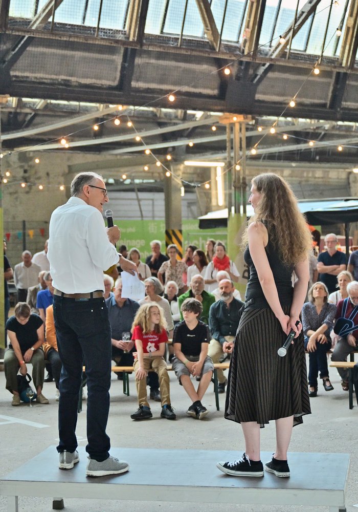 Zwei Personen, die in der festlich geschmückten Shedhalle vor vielen Menschen eine Rede halten.
